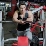 A young man resting in a gym while using a mobile phone beside weightlifting equipment.