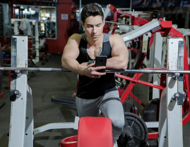 A young man resting in a gym while using a mobile phone beside weightlifting equipment.