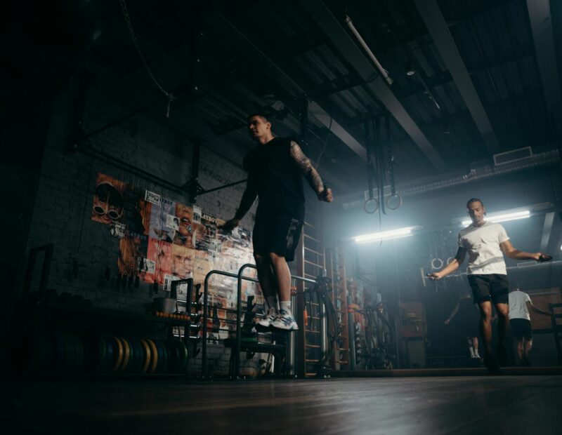 Two men actively skipping jump ropes in a dimly lit gym environment.